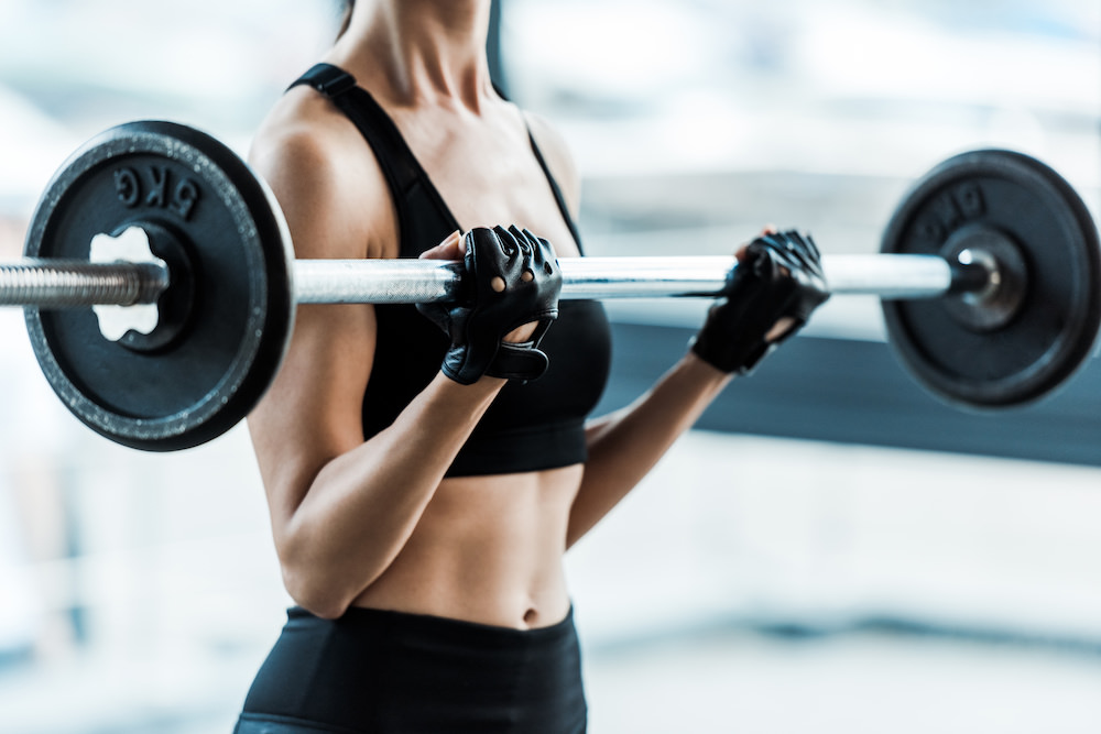cropped view of young woman in sportswear exercising with barbell in gym